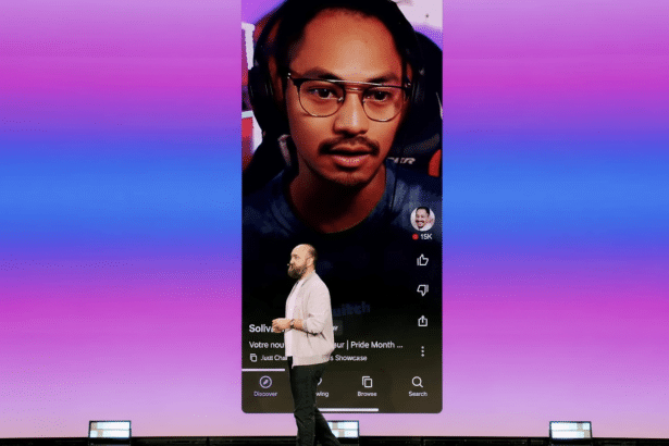 A man standing on a stage in front of a giant screen displaying a vertical video of another man wearing headphones. The screen has a purple and blue gradient background.