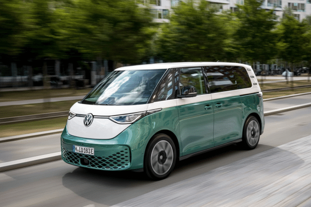 A two -tone white and teal Volkswagen ID. Buzz electric van driving on a road with trees and buildings in the background.