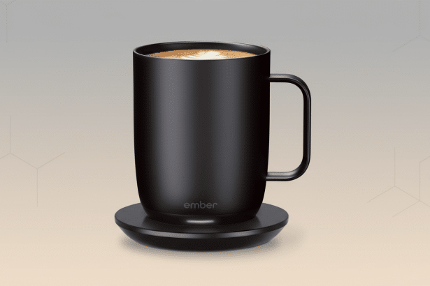 A matte black Ember smart mug with coffee , featuring a white latte art design , sitting on its black charging coaster. The background is a soft gradient from light beige to grey with subtle hexagonal patterns .