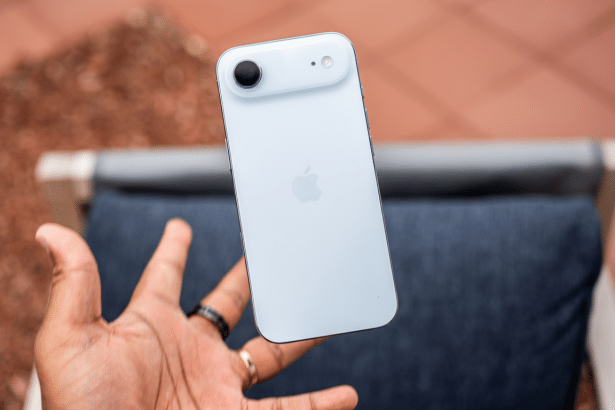A hand holding a white iPhone 15 vertically, showcasing its back with the Apple logo, camera, and flash, against a slightly blurred outdoor background