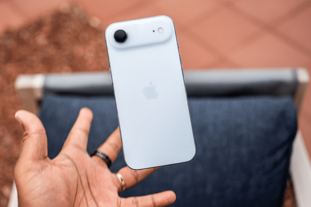 A hand holding a white iPhone 15 vertically, showcasing the back with its camera module and Apple logo, against a slightly blurred outdoor background