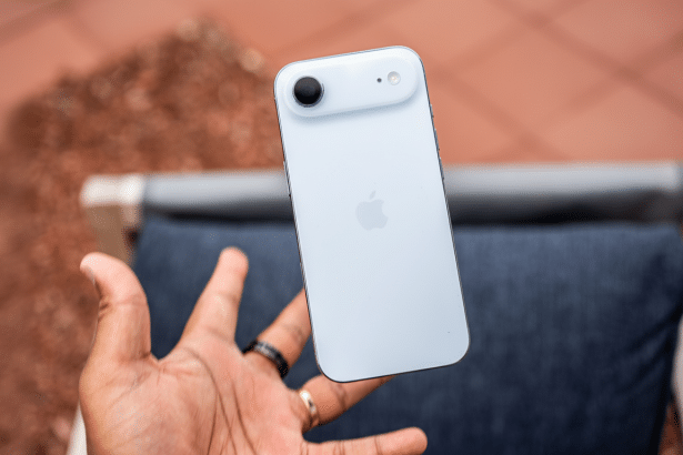 A hand holds a white iPhone, showcasing its back camera and Apple logo, with an outdoor background.