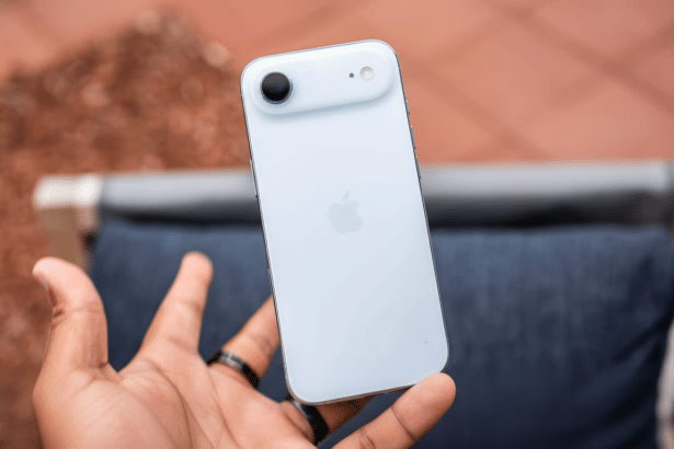 A hand holds a white iPhone, showcasing its back camera and the Apple logo. The background is a blurry outdoor setting with reddish -brown pavement an
