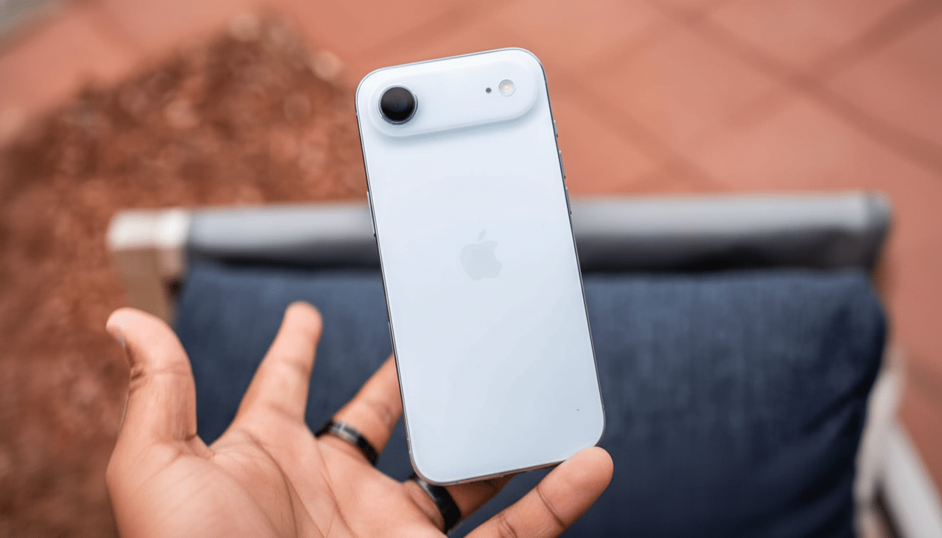 A hand holds a white iPhone, showcasing its back camera and the Apple logo. The background is a blurry outdoor setting with reddish -brown pavement an