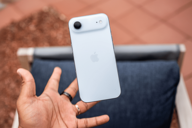 A hand holding a white iPhone with a single camera lens and an Apple logo on the back , against a blurred outdoor background.