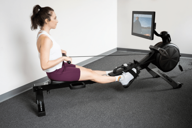 A woman in athletic wear sits on a black NordicTrack RW 900 rower , gripping the handle and extending her legs . A screen attached to the rower displays a scenic outdoor view. Filename : womannord ictrackrow er.png