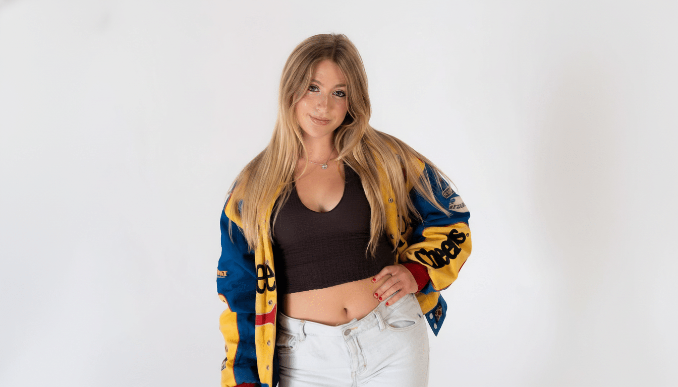 A young woman with long blonde hair, wearing a racing jacket, black crop top, and light jeans, posing against a white background.