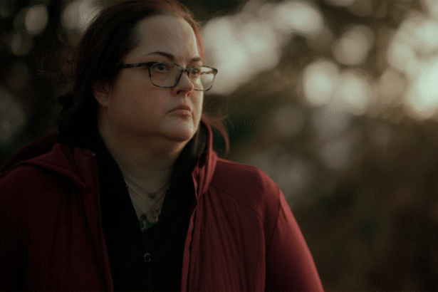 A woman with glasses and a red jacket looks to her right with a serious expression, against a bokeh background of soft outdoor light. Filename : womanred jacketoutdoor portrait .png