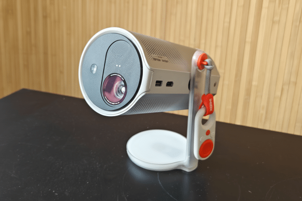 A professional shot of a white and silver portable projector with a red strap , resting on a dark surface with a light wooden background , presented in a 16:9 aspect ratio.