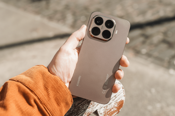 A hand in an orange corduroy sleeve holds a metallic brown Xiaomi smartphone , featuring a prominent quad-camera array with Leica branding, against a blurred outdoor background.