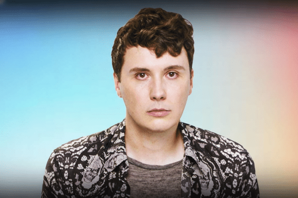 A young man with brown hair and a patterned shirt looking directly at the viewer , against a soft gradient background.