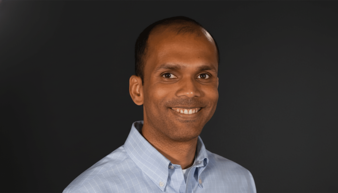 A professional headshot of a man with dark skin and short dark hair, wearing a light blue collared shirt, smiling against a dark gray background.