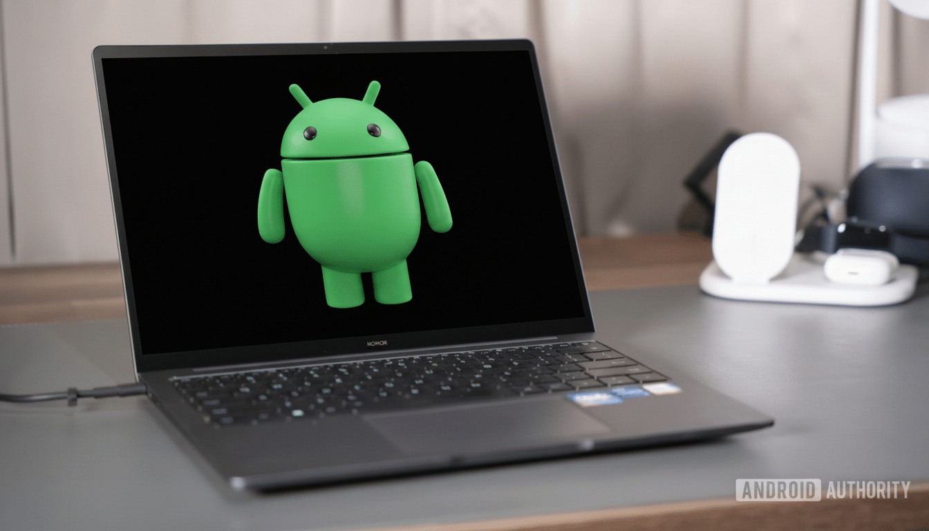 A laptop displaying the Android robot logo on its screen, set on a desk with various tech accessories in the background.