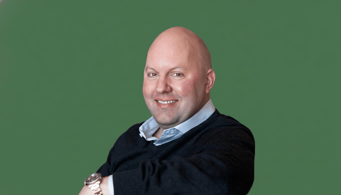 A bald man with a light complexion, wearing a dark blue sweater over a light blue collared shirt, smiles at the camera. He has a watch on his left wrist and is positioned against a solid green background.