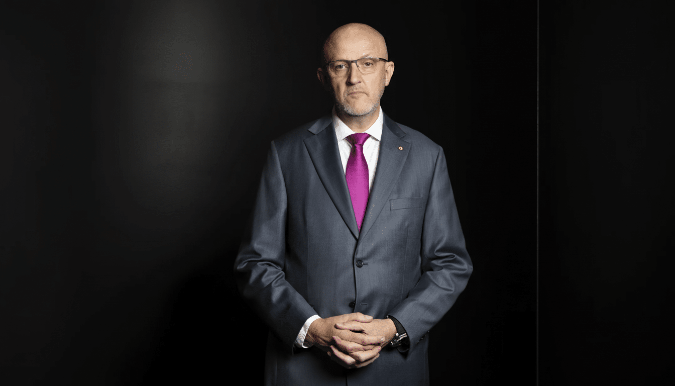 A bald man with glasses and a beard, wearing a grey suit and a bright pink tie, stands against a dark background with his hands clasped in front of him.