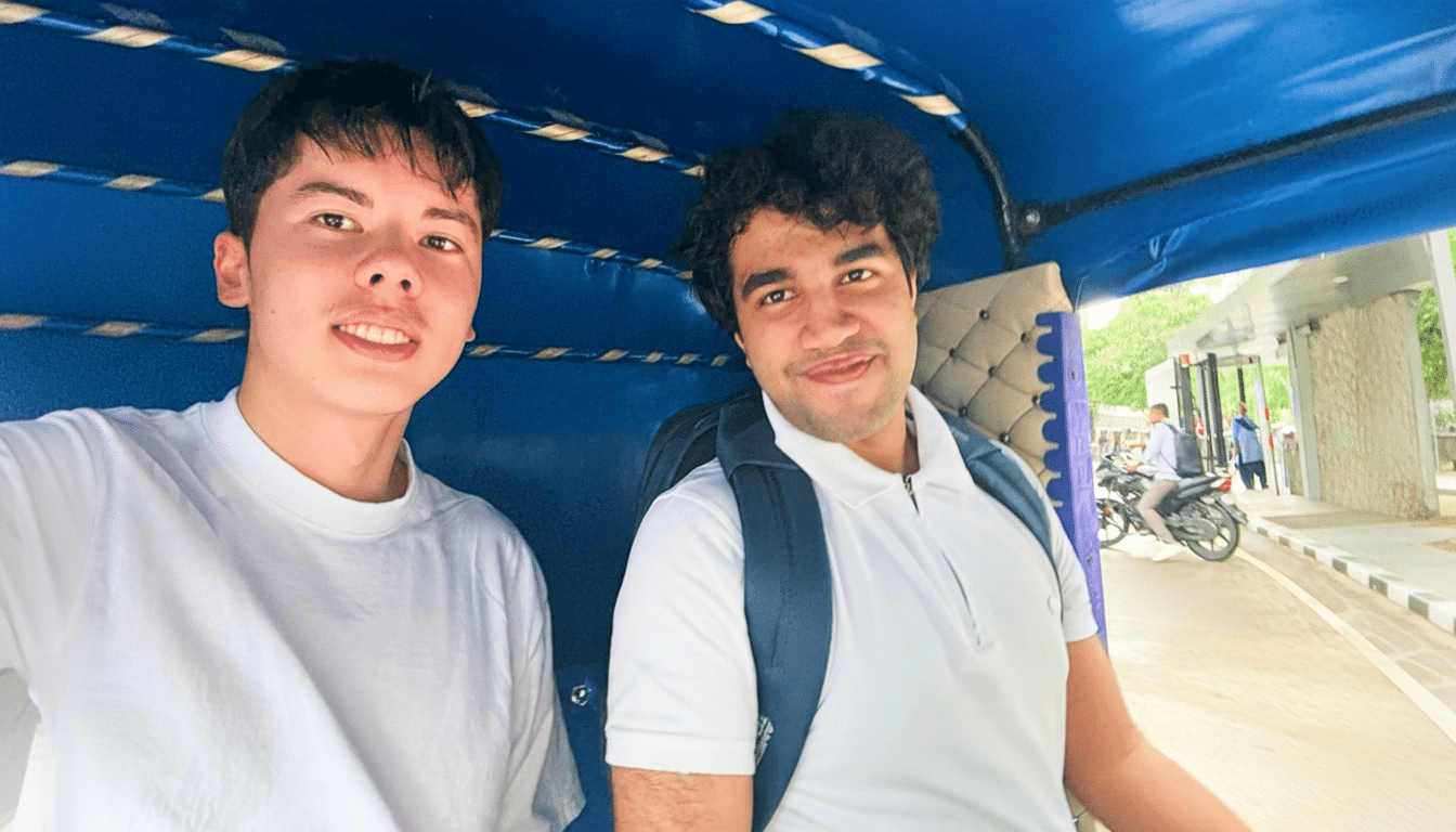 Two young men, one with dark hair and a white t-shirt, and the other with curly dark hair and a white polo shirt with a backpack, are sitting inside a blue vehicle, possibly a tuk-tuk, smiling at the camera. The background shows a street scene with a motorcycle and people.