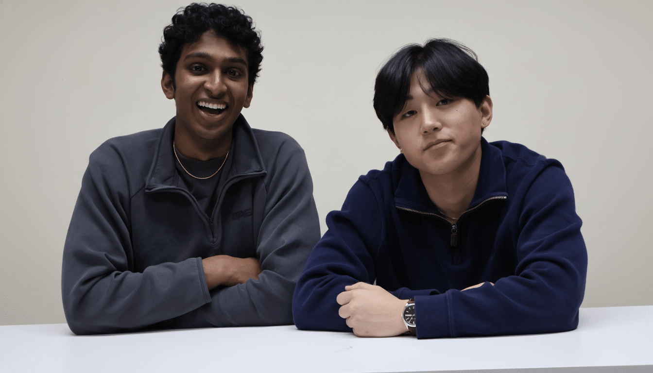 Two young men, one with dark curly hair and a wide smile, and the other with straight dark hair and a neutral expression, are seated at a white table against a light background.