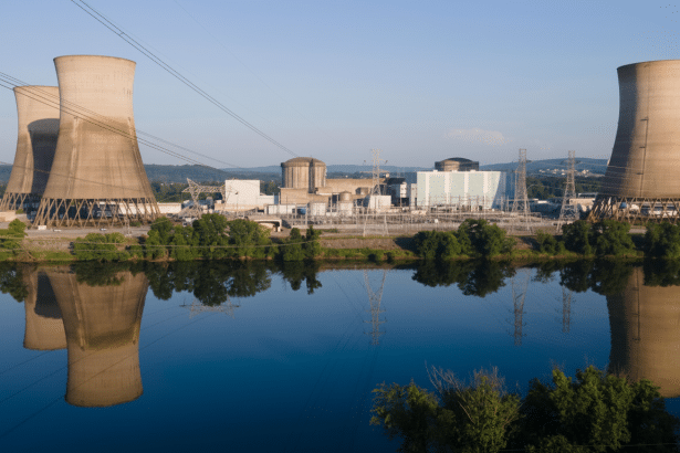 A nuclear power plant with three cooling towers reflected in the calm blue water of a river, under a clear sky.