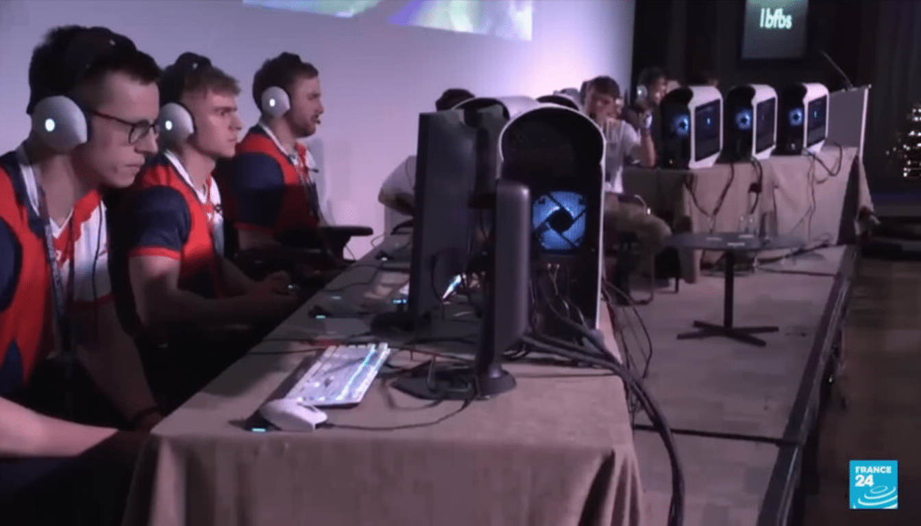 A group of young men wearing headphones and matching shirts are seated at a long table with computers, appearing to be engaged in an esports competition.