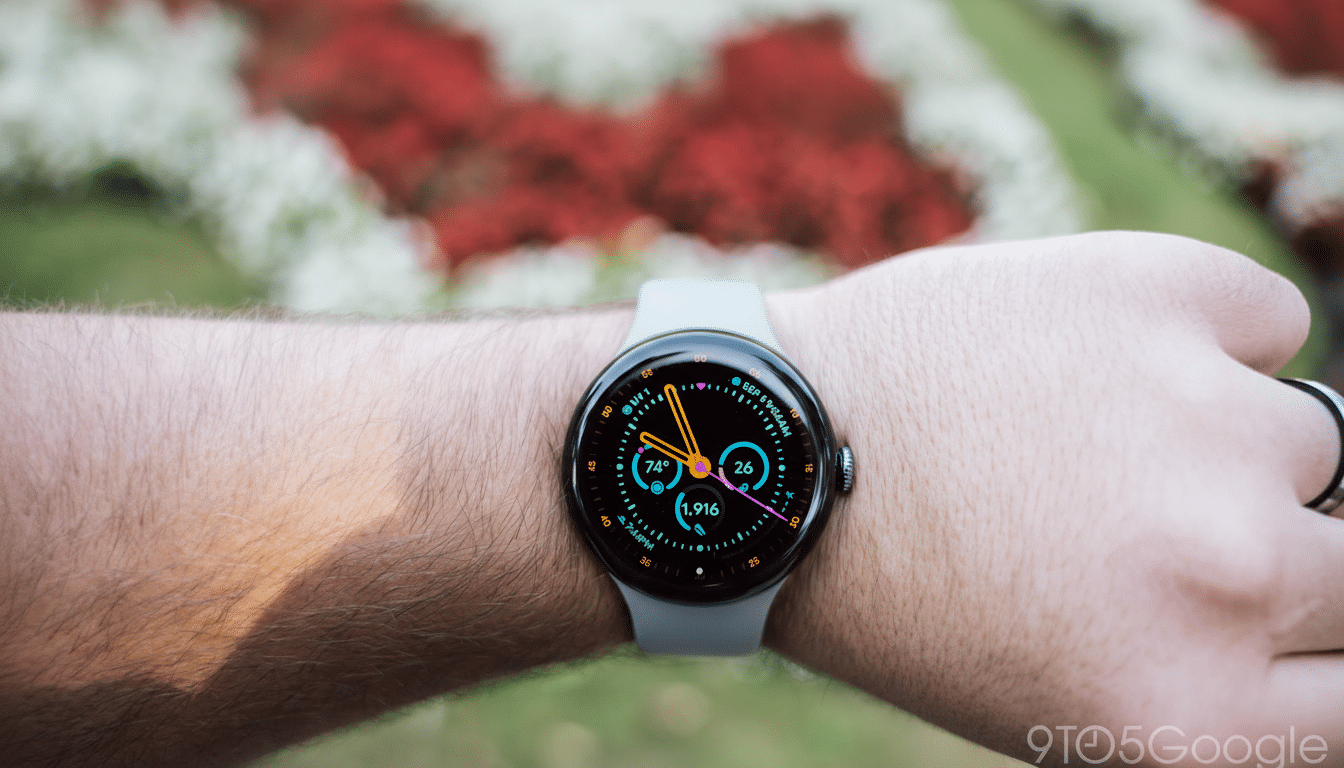 A close-up of a persons wrist wearing a Google Pixel Watch with a colorful watch face displaying time, weather, and activity data, set against a blurred outdoor background of red and white flowers.