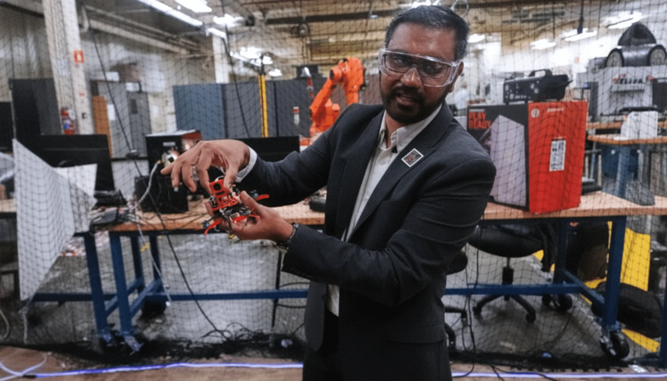 A man in safety glasses and a suit holds a small drone in a workshop setting.