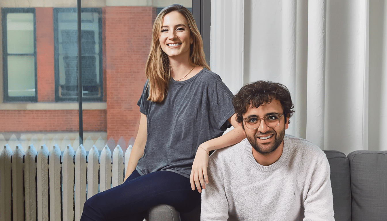A smiling woman with blonde hair in a grey t-shirt and dark pants sits on a grey armrest, leaning on a smiling man with dark curly hair and glasses in a light-colored sweater, who is seated on a grey couch. They are in a room with a large window showing a brick building outside.