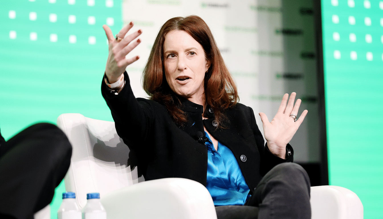 A woman with reddish-brown hair, wearing a black jacket and a blue top, sits in a white chair with her hands gesturing as she speaks.