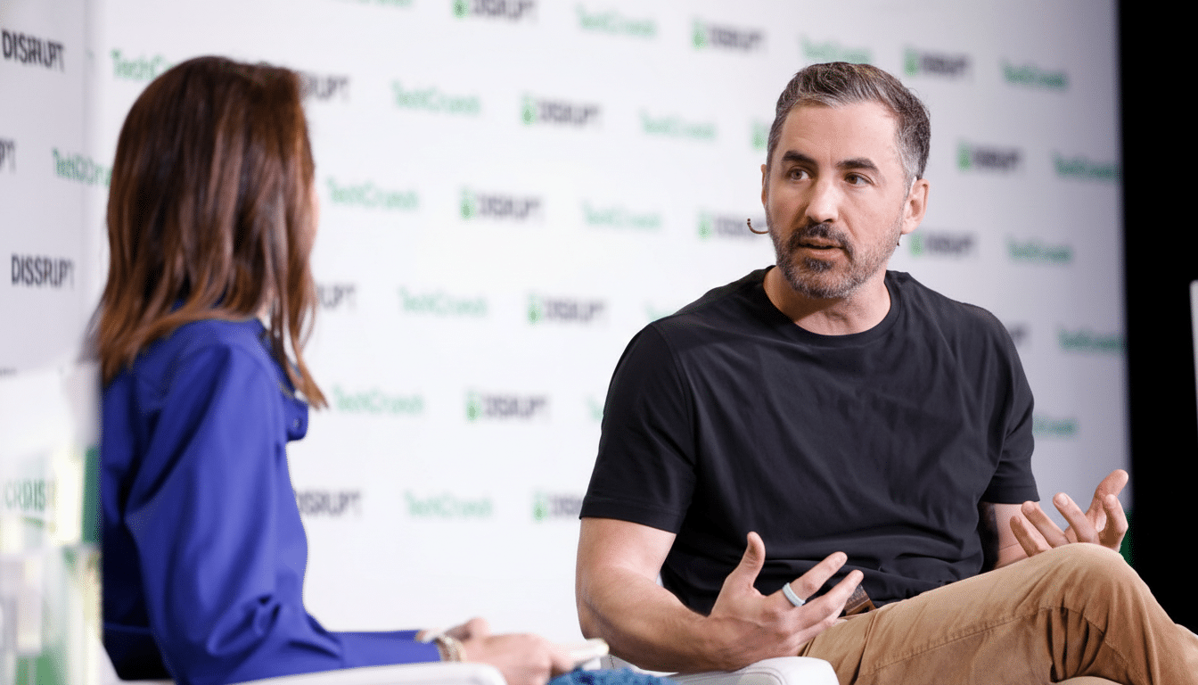 A man in a black t-shirt and tan pants speaking animatedly to a woman in a blue shirt, both seated on white chairs against a white background with green TechCrunch Disrupt logos.