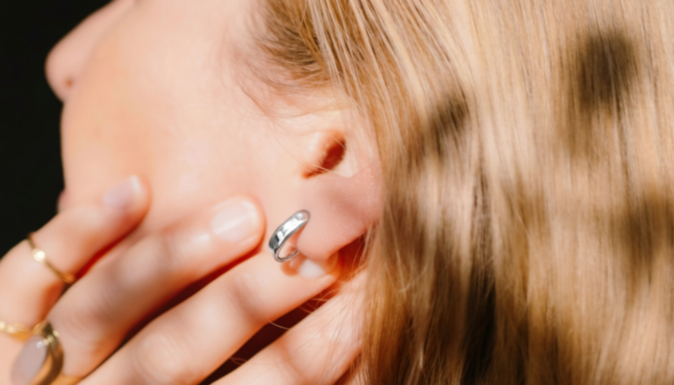 A close-up of a persons ear wearing a silver hoop earring, with blonde hair and a hand partially visible.