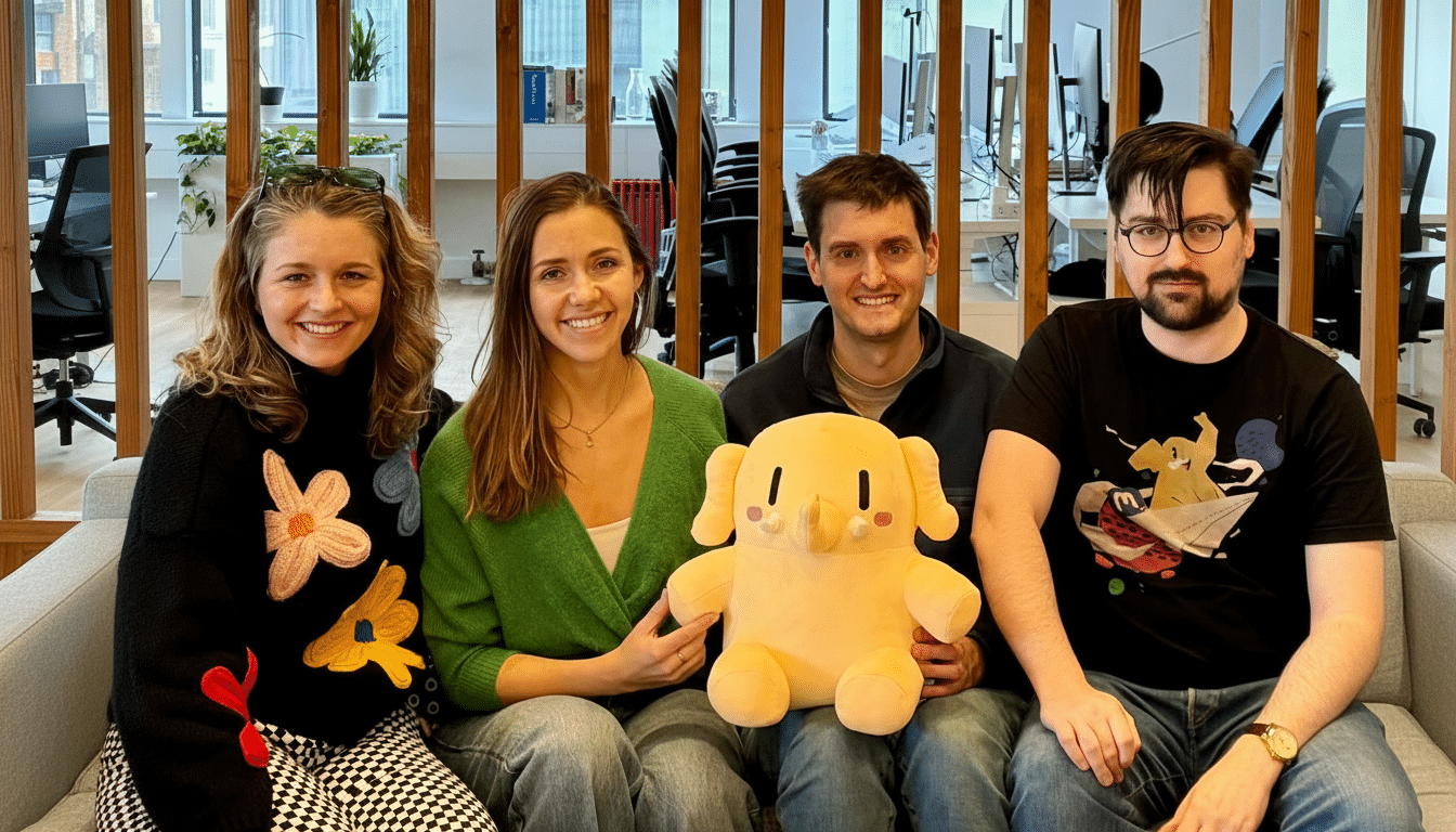 Four people, two women and two men, are sitting on a couch in an office setting, smiling at the camera. The man in the middle is holding a large, yellow elephant plush toy.