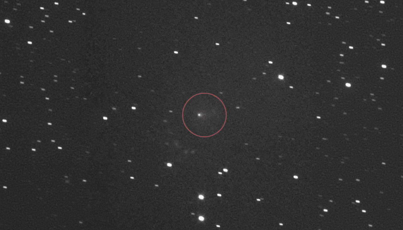 A black and white astronomical image showing a field of stars, with a faint, slightly elongated object circled in red at the center.