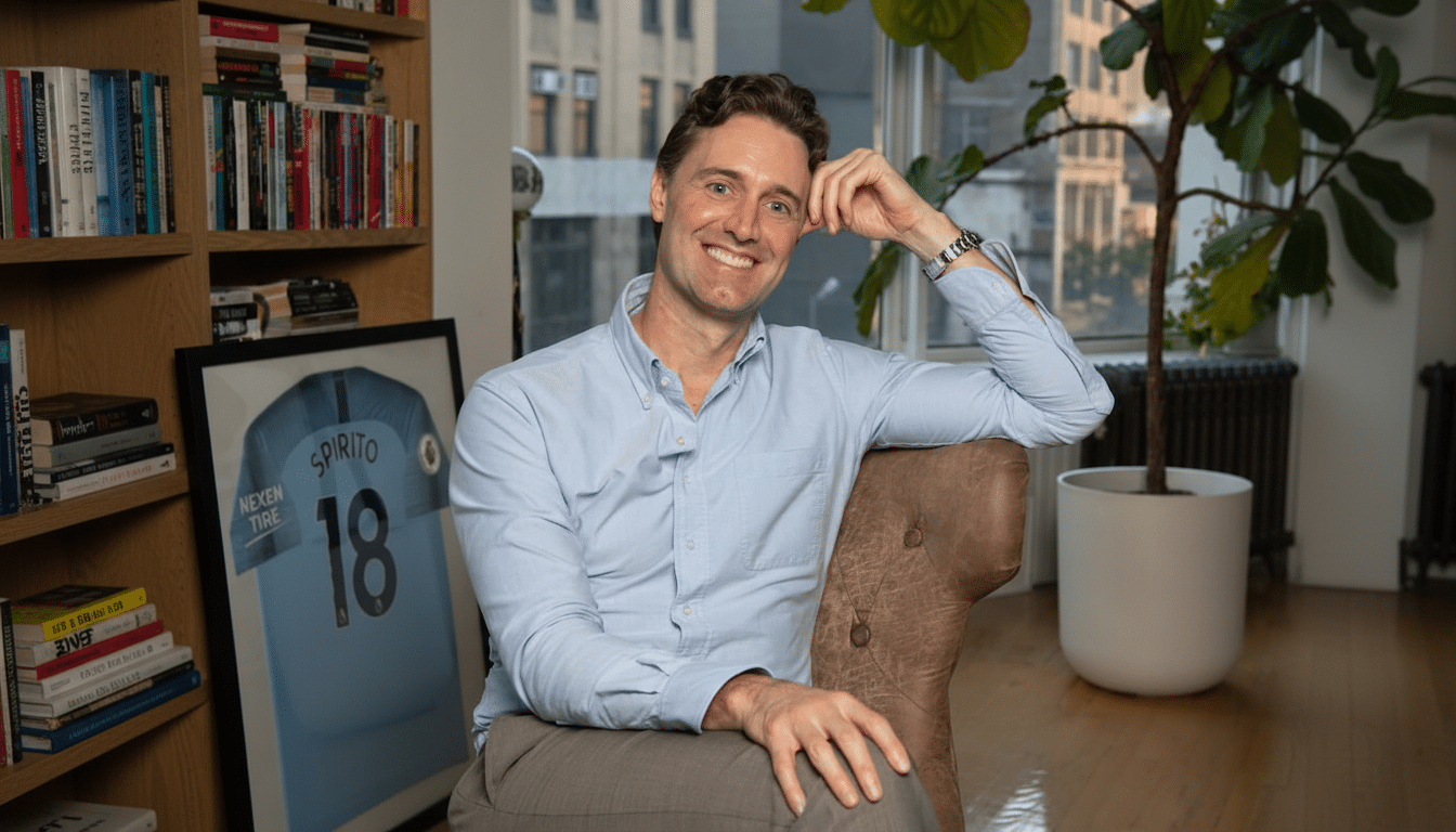 A man in a light blue shirt and grey pants smiles at the camera while sitting in a brown leather chair. He is in a room with a bookshelf, a framed soccer jersey, and a large plant.