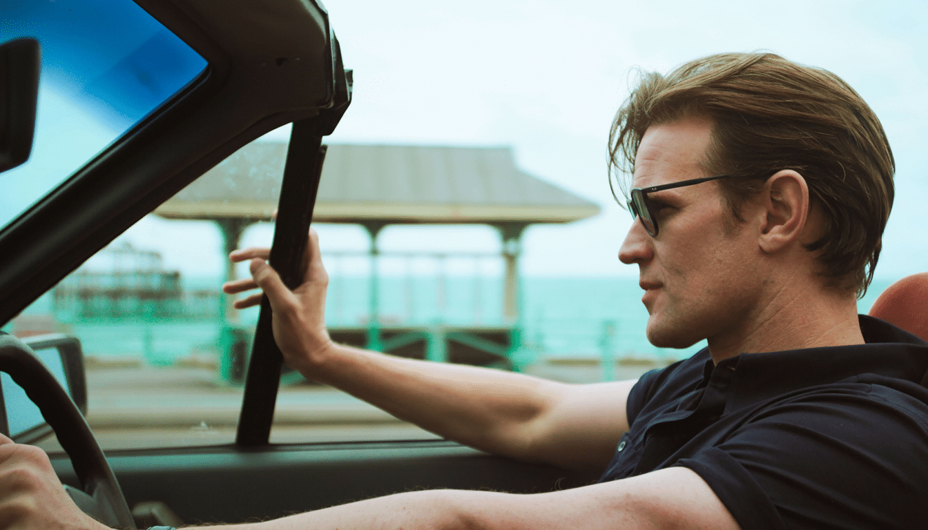 A man with sunglasses driving a convertible car by the sea.