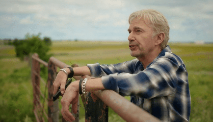 A man with light hair and a blue plaid shirt leans on a fence in a field, looking into the distance.
