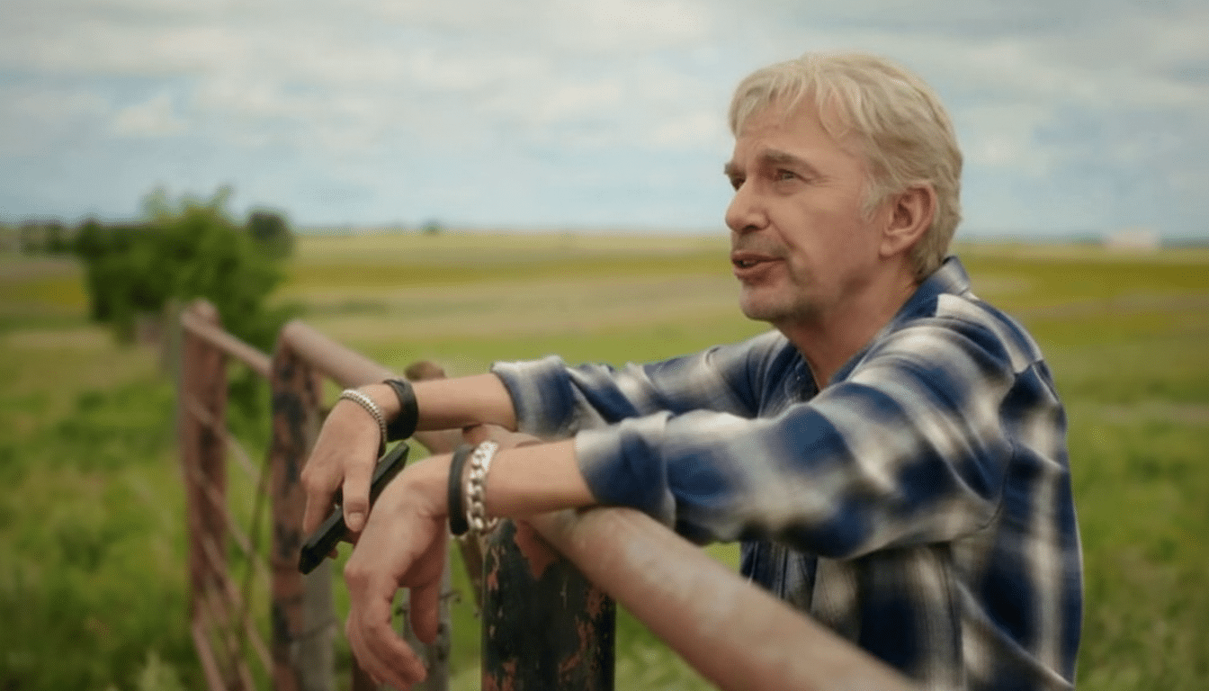 A man with light hair and a blue plaid shirt leans on a fence in a field, looking into the distance.