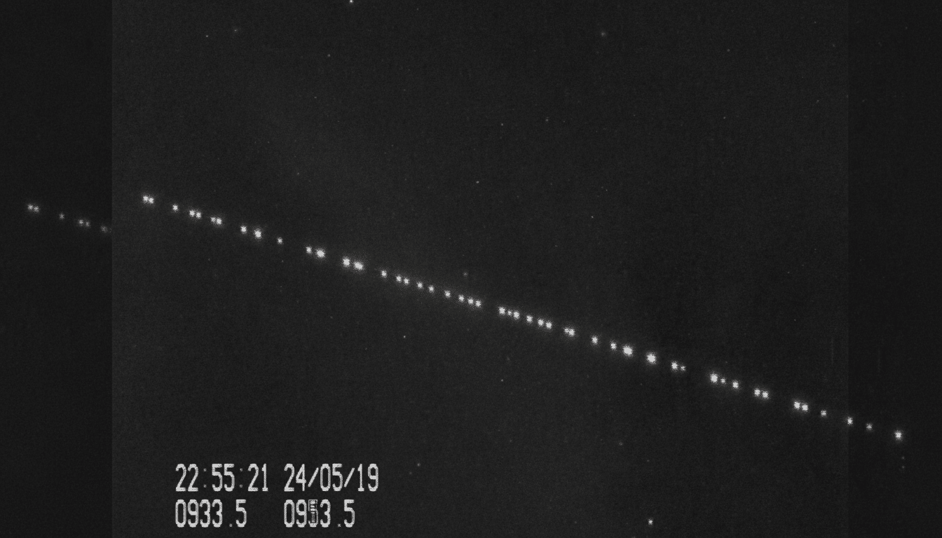 A black and white image showing a diagonal line of evenly spaced bright dots, likely satellites, against a dark background with faint stars. A timestamp and numerical data are visible in the bottom left corner.