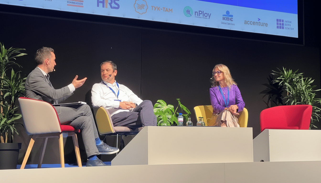 Three people on a stage, two men and one woman, engaged in a panel discussion with sponsor logos displayed on a screen behind them.
