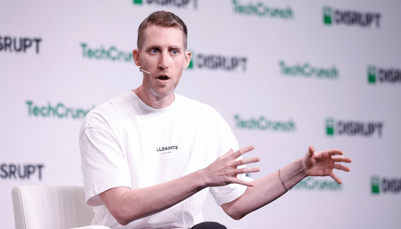 A man with short hair and a white t-shirt speaking at an event with TechCrunch Disrupt logos in the background.