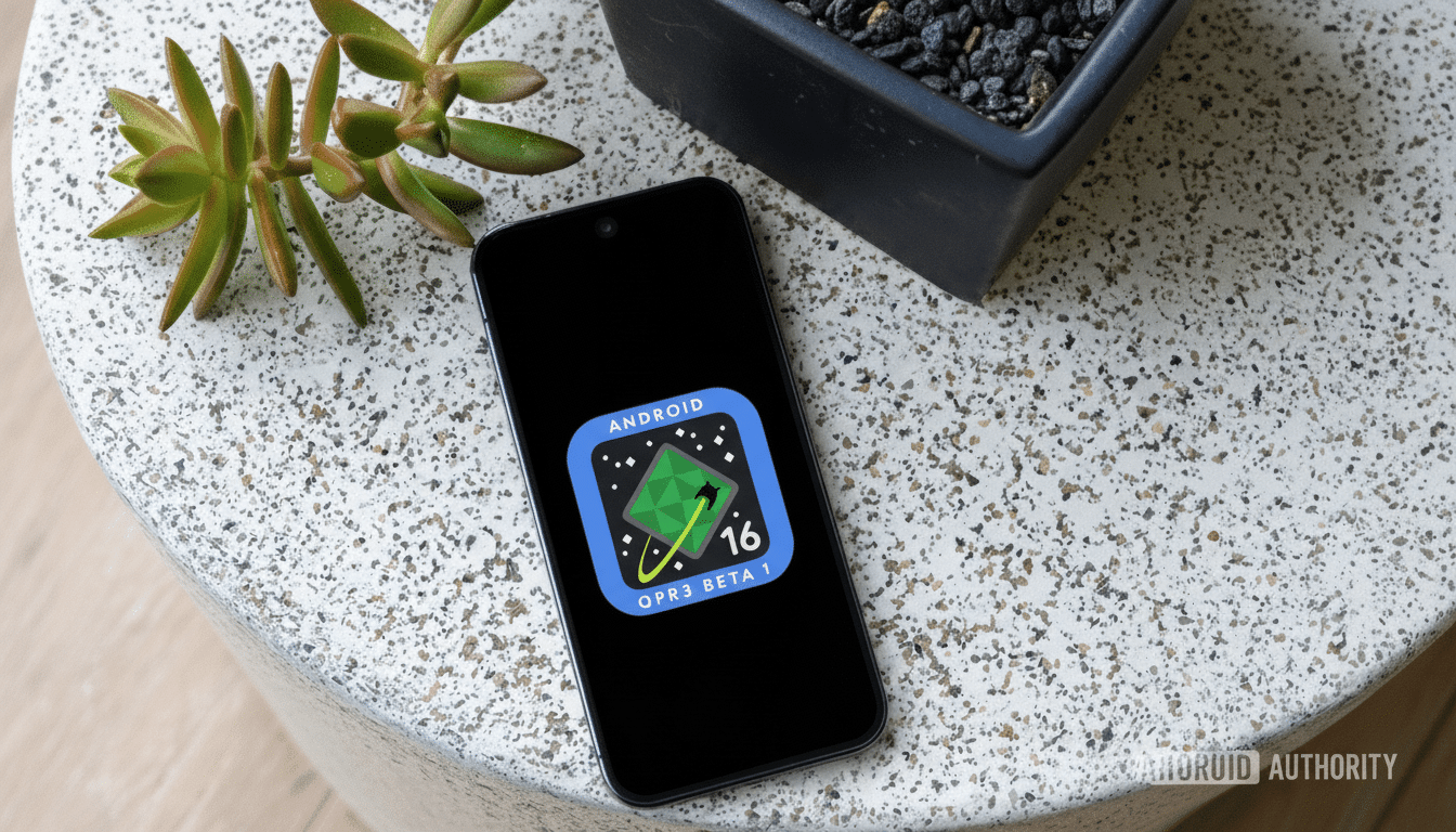 A smartphone displaying the Android 16 QPR3 Beta 1 logo on its screen, placed on a speckled white surface next to a small green plant and a black pot with dark pebbles.