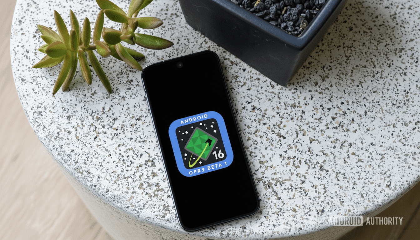 A smartphone displaying the Android 16 OPR3 Beta 1 logo on its screen, placed on a speckled white surface next to a small green plant and a dark pot with pebbles.