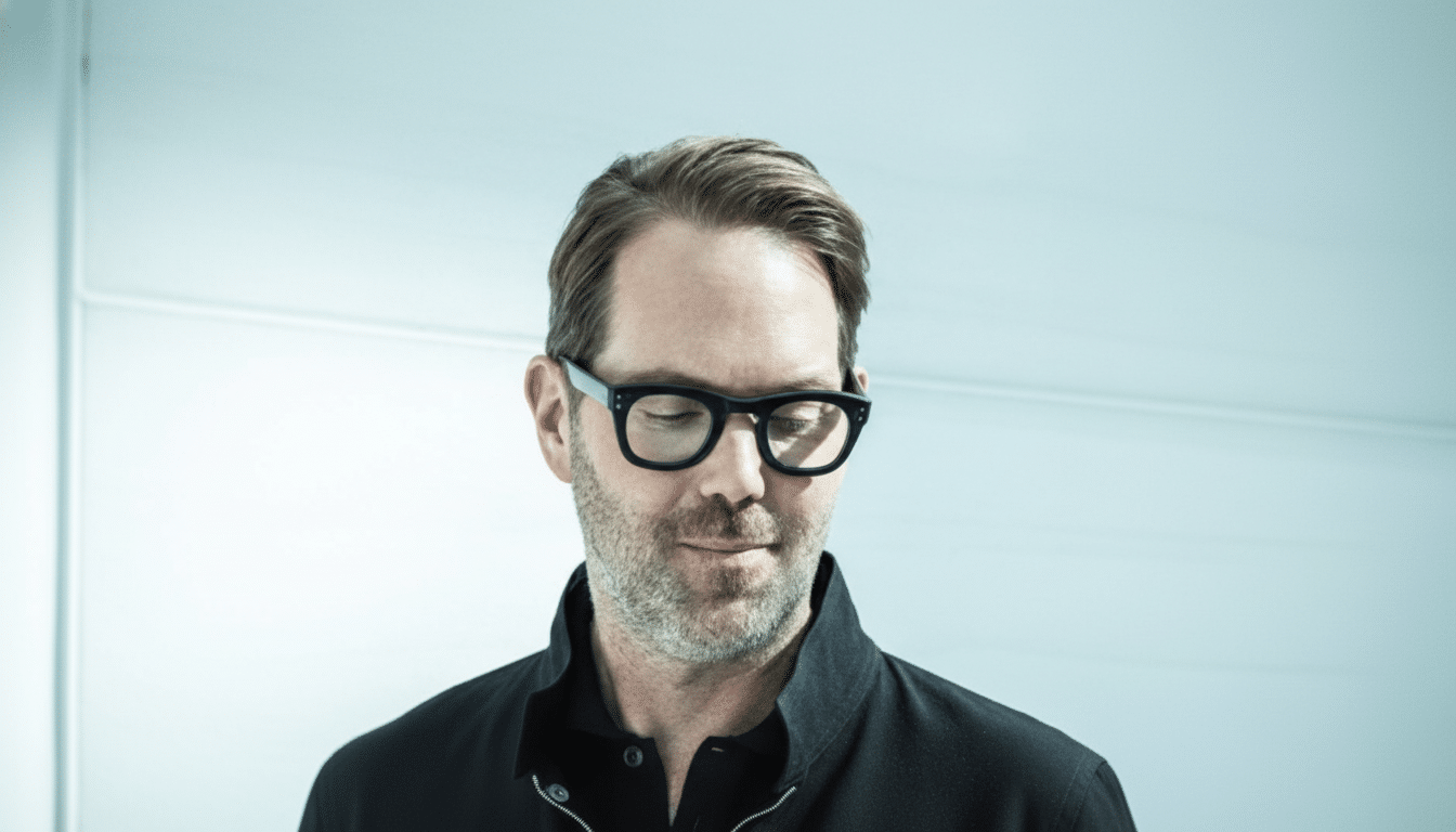 A man with glasses and a beard, wearing a black jacket, looking down.