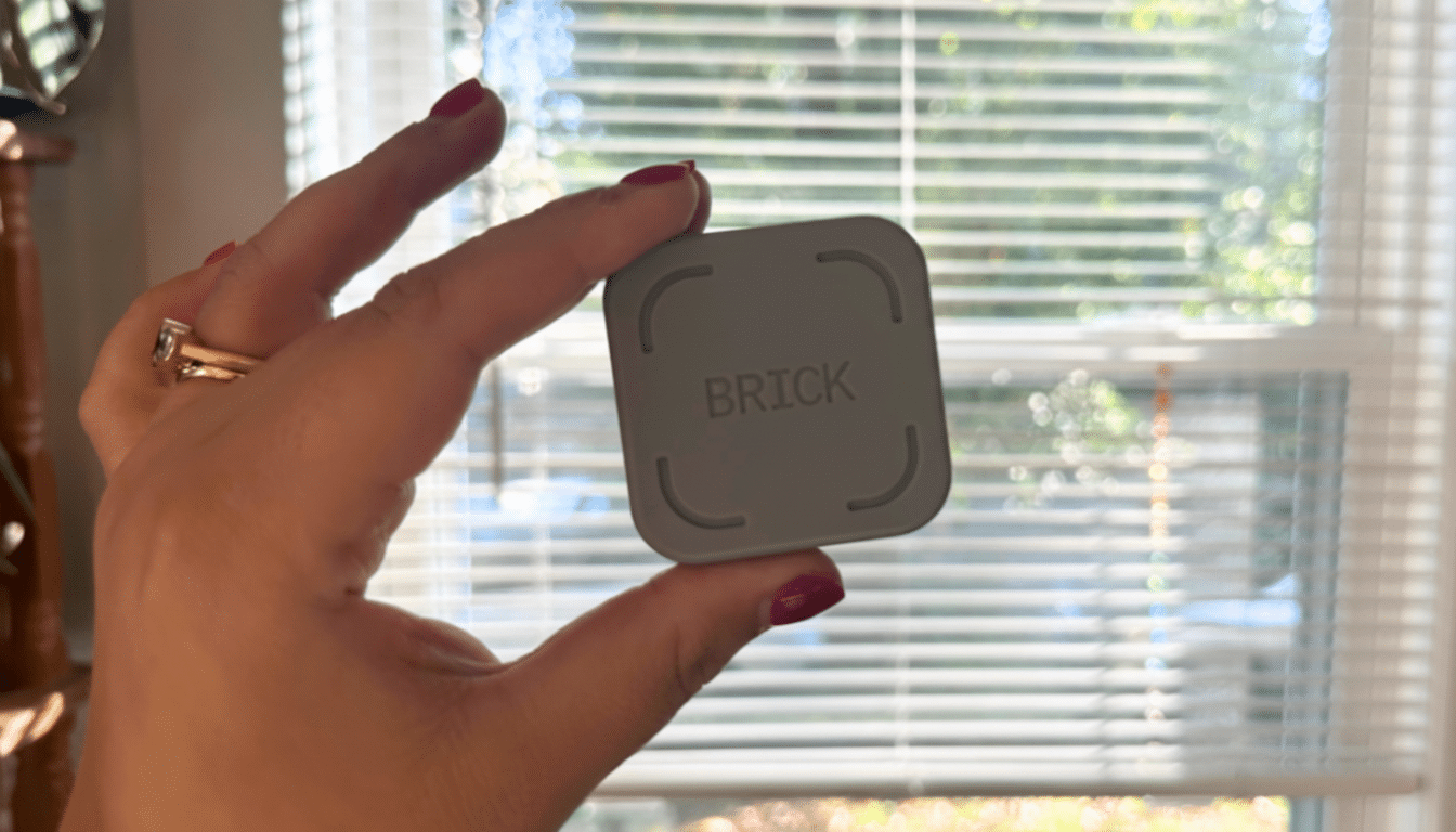 A hand holding a gray square device with the word BRICK on it, against a blurred background of a window with blinds.