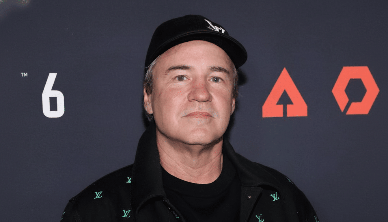 A man wearing a black baseball cap and a black jacket with green logos, standing in front of a dark background with a white 6 and an orange triangular logo.