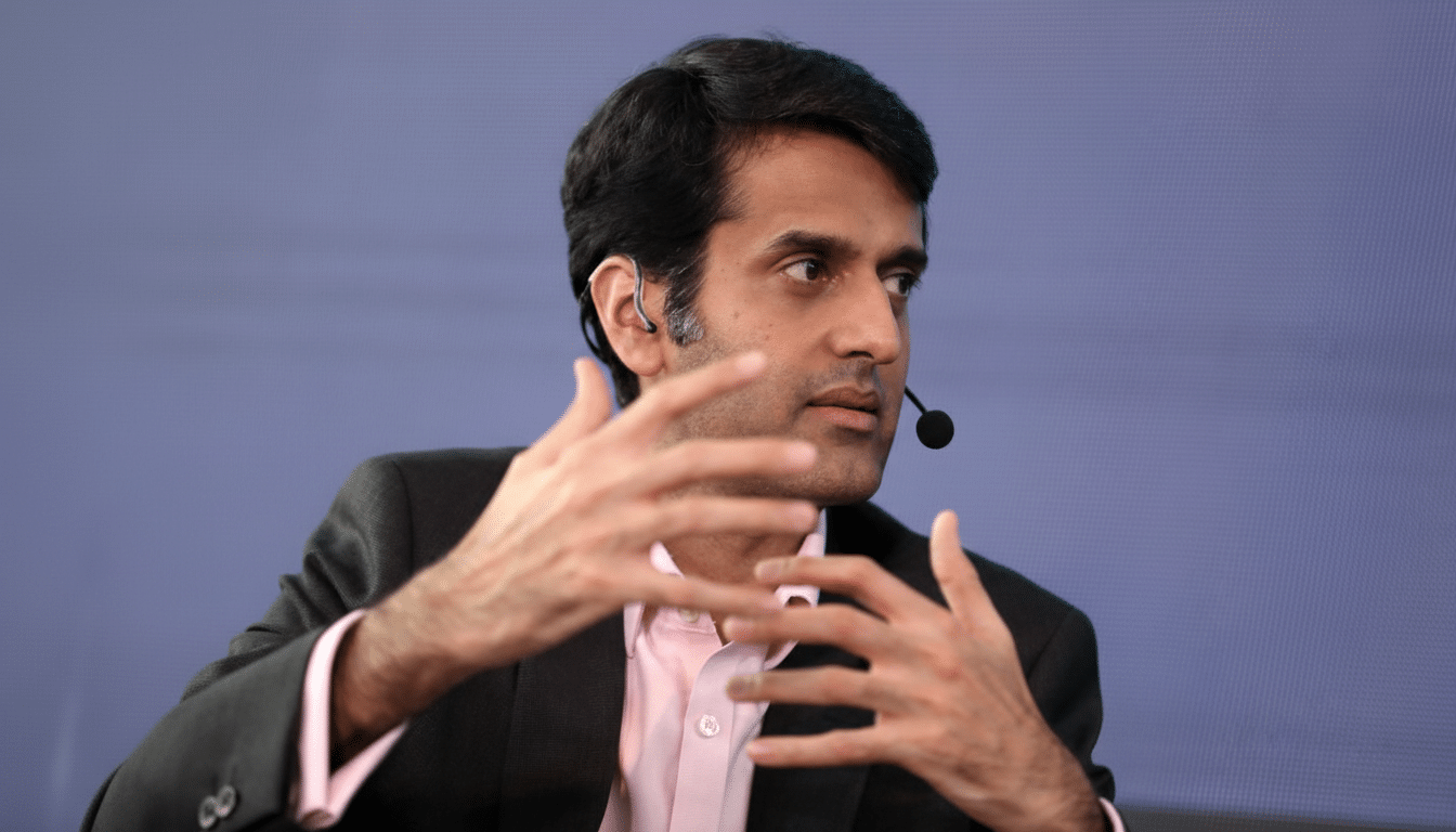 A man in a suit and pink shirt, wearing a headset microphone, gestures with both hands while speaking.