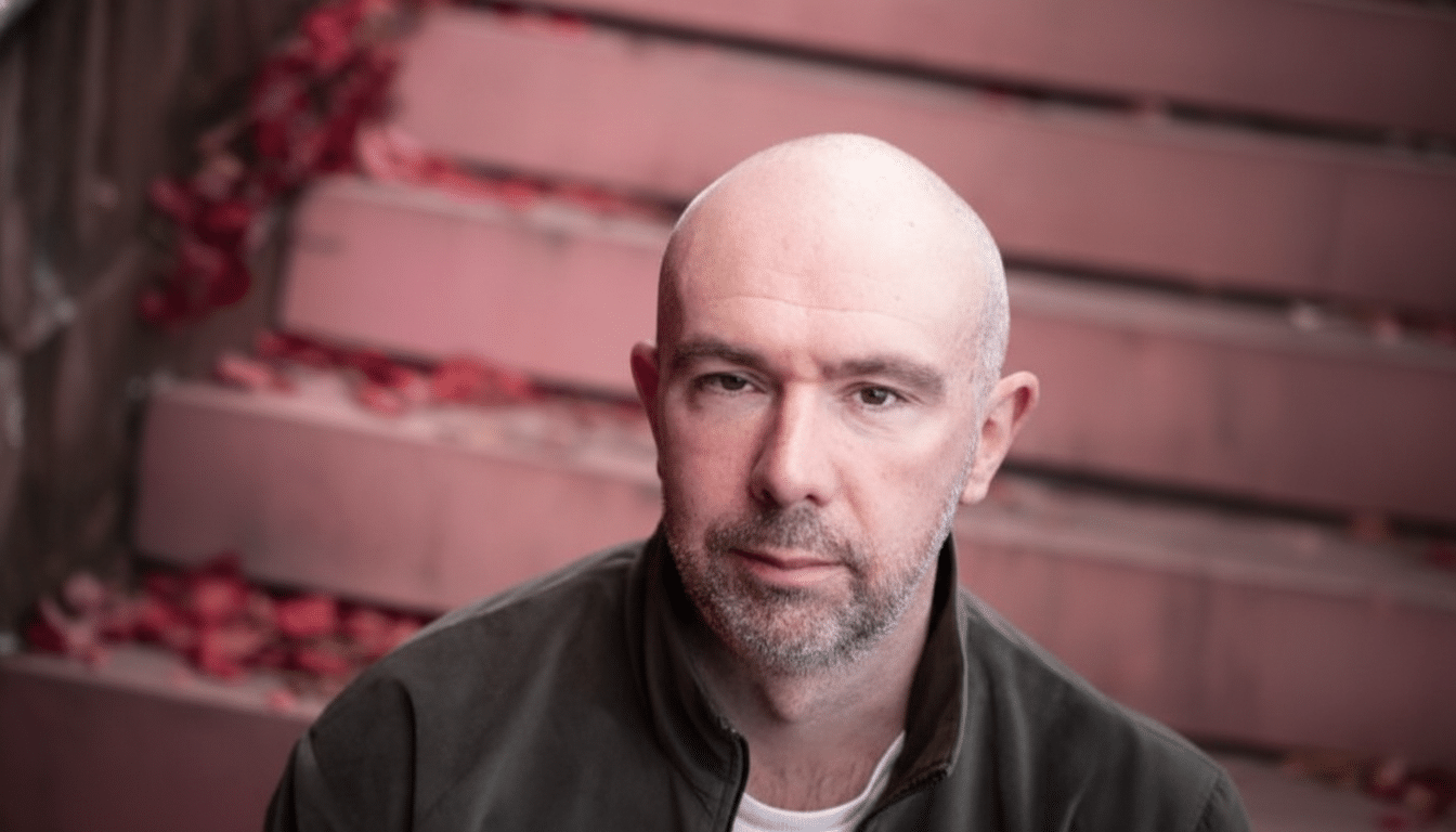 A bald man with a beard looking directly at the camera, with a blurred background of pink steps and scattered red petals.