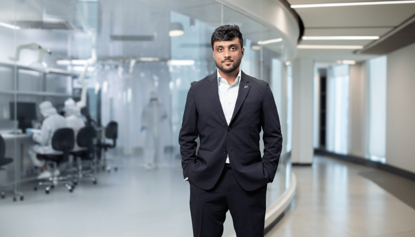 A man in a suit stands in a modern office setting with a laboratory visible in the background.