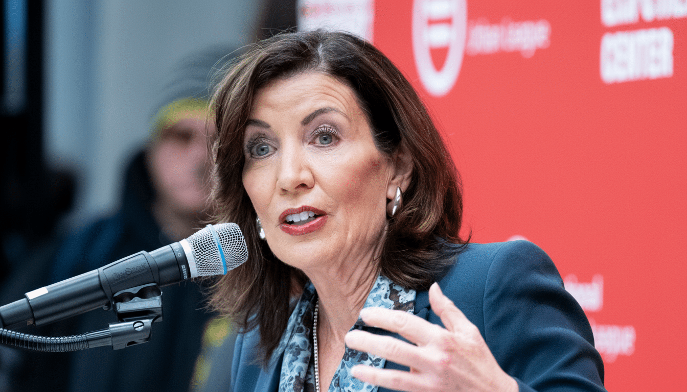 A woman with dark hair and a blue blazer speaks into a microphone, with a red background behind her.