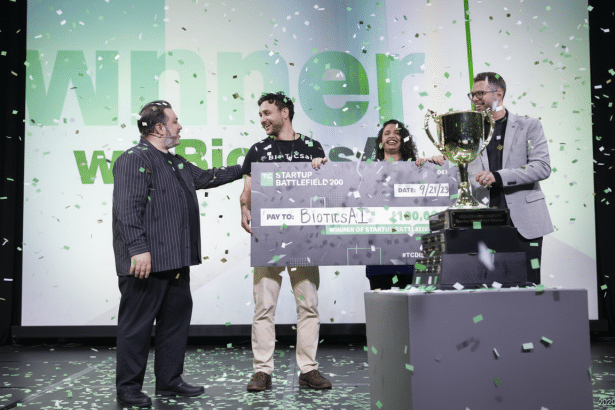 Four people on a stage, with one man on the left handing a large check to another man in the center, who is holding it with a woman. A fourth man stands to the right of a large trophy. Green confetti falls around them.