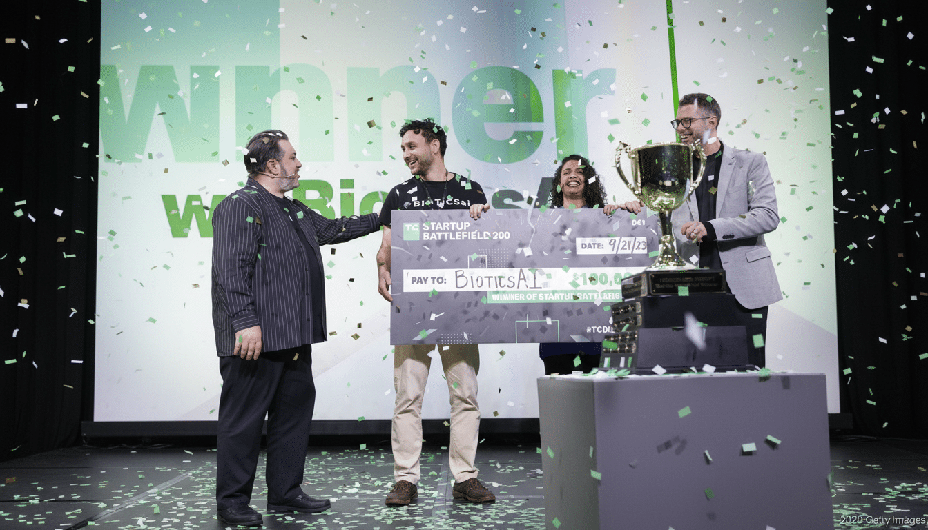 Four people on a stage, with one man on the left handing a large check to another man in the center, who is holding it with a woman. A fourth man stands to the right of a large trophy. Green confetti falls around them.