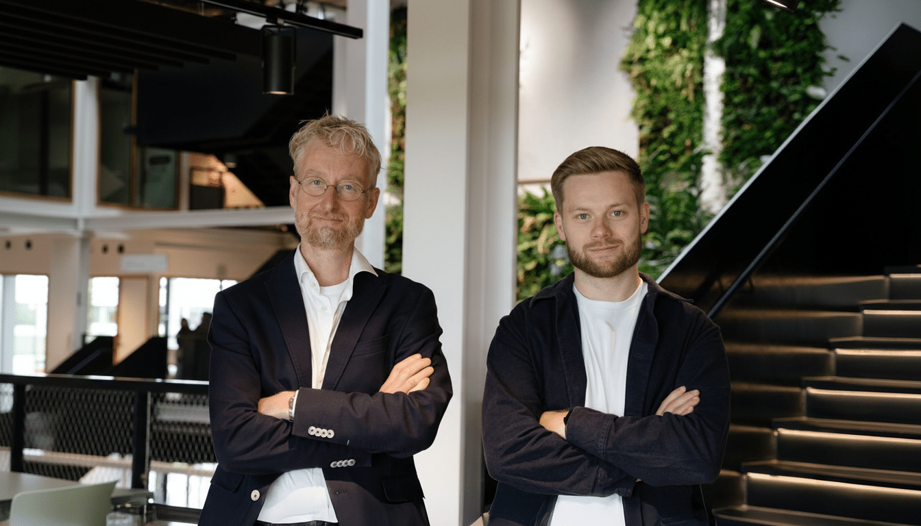 Two men standing side-by-side with their arms crossed, looking at the camera. The man on the left has glasses and a beard, wearing a dark suit jacket over a white shirt. The man on the right has a beard and is wearing a dark jacket over a white t-shirt. They are in an indoor setting with a modern design, including a green wall with plants and a staircase in the background.